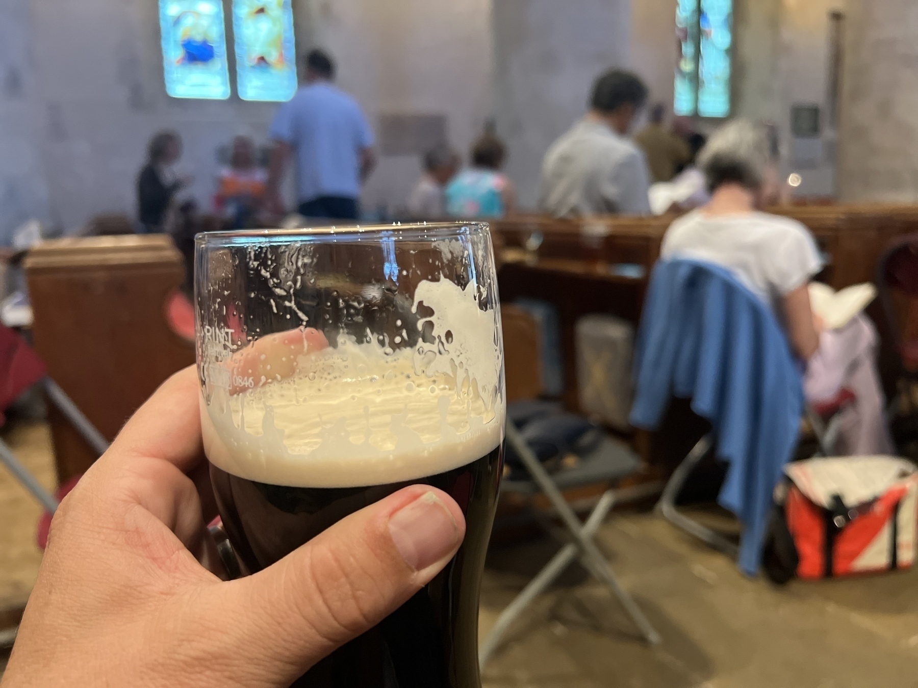 A person is holding a pint of stout inside a church where other people are seated.